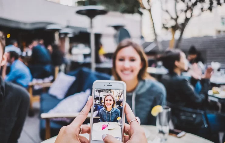 Person, die in einem Straßencafé ein Foto einer lächelnden Frau mit einem Smartphone macht.