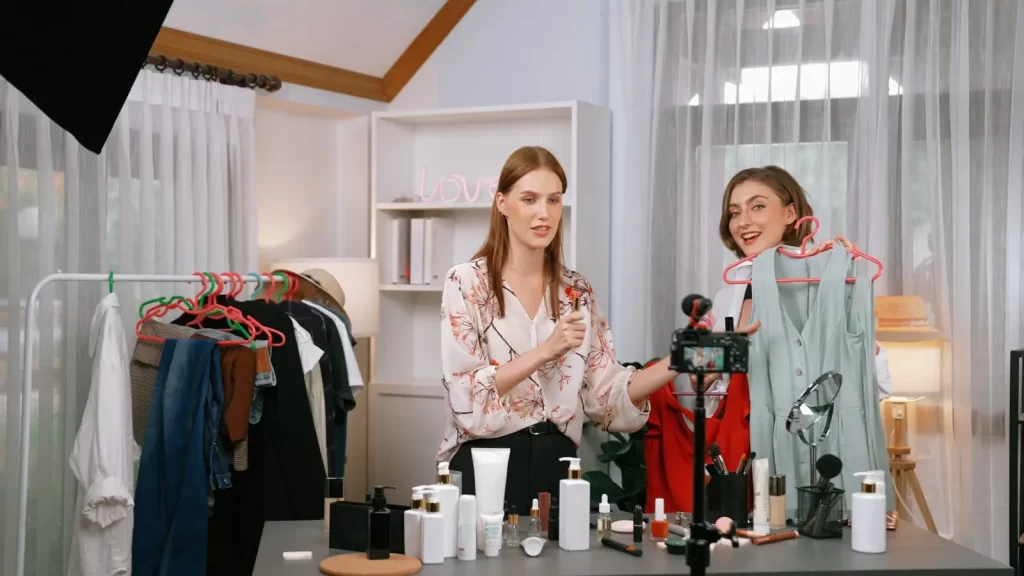 Zwei Frauen nehmen in einem gut beleuchteten Raum mit Kleidung und Make-up-Produkten einen Mode- und Beauty-Videoblog auf.