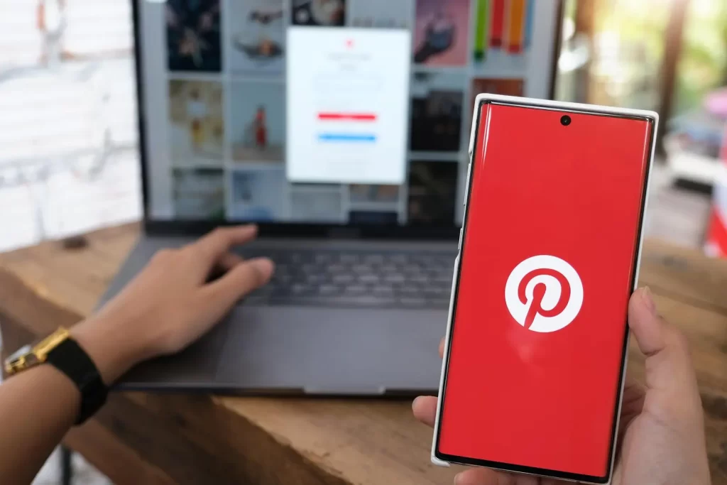 Person, die ein Smartphone mit dem Pinterest-Logo vor einem Laptop hält, auf dem der Pinterest-Anmeldebildschirm angezeigt wird.