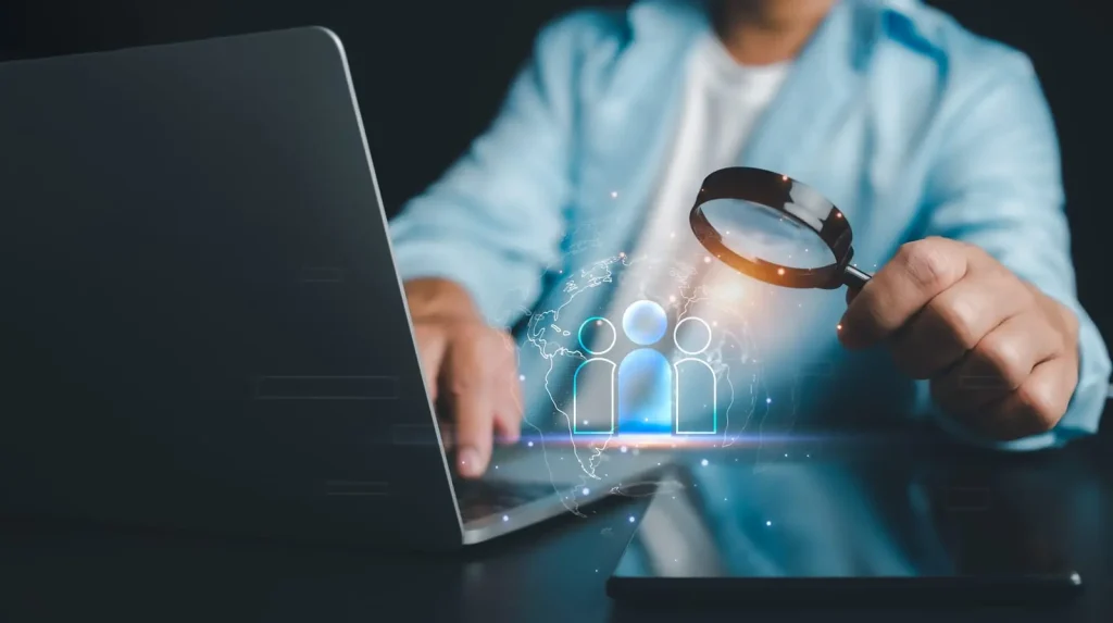 Person, die eine Lupe über einen Laptop-Bildschirm hält, auf dem digitale Personensymbole angezeigt werden, die eine Anwerbung oder Suche symbolisieren.