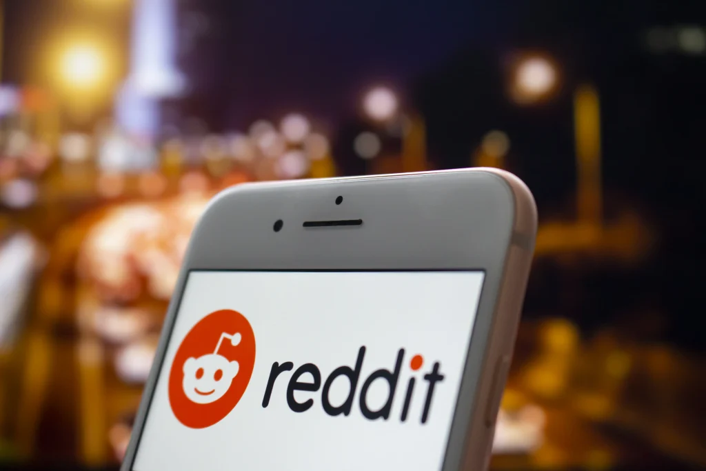 Nahaufnahme eines Smartphones, auf dem das Logo der Reddit-App mit einer unscharfen nächtlichen Stadtlandschaft im Hintergrund angezeigt wird.