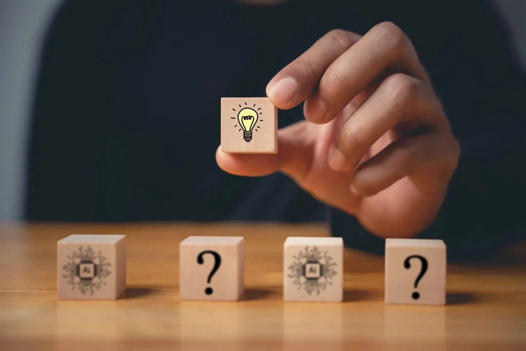 Eine Hand platziert einen Holzblock mit einem Glühbirnensymbol zwischen Blöcken mit Fragezeichen und KI-Symbolen auf einer Holzoberfläche.
