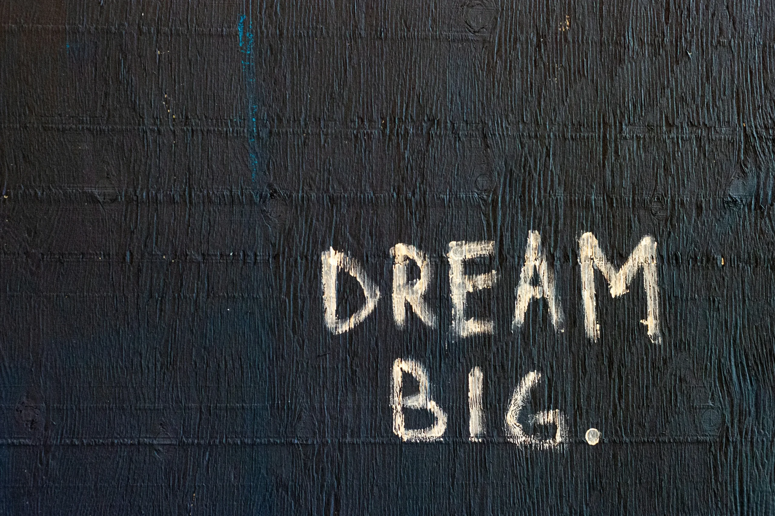 Das Bild zeigt den Satz „Dream Big“ in Weiß auf einem dunklen strukturierten Hintergrund.