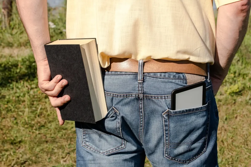Eine Person steht im Freien, hält in einer Hand ein dickes Buch und in der Gesäßtasche ihrer Jeans einen E-Reader.