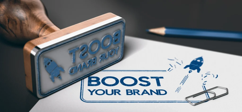 Auf einem Stempel und Papier ist der Satz „Boost Your Brand“ mit einem Raketensymbol zu sehen. Ein Bleistift und eine Büroklammer liegen daneben.