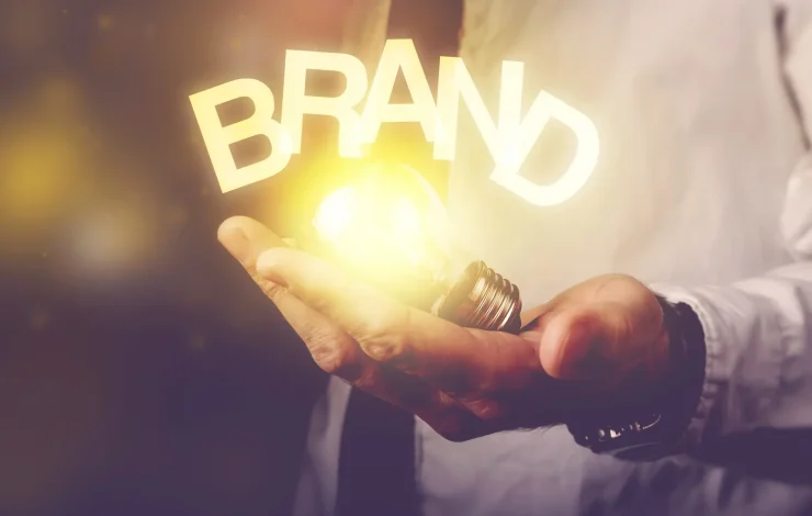 Eine Person hält eine leuchtende Glühbirne, über der das Wort „MARKE“ schwebt, was das Konzept der Markeninnovation symbolisiert.