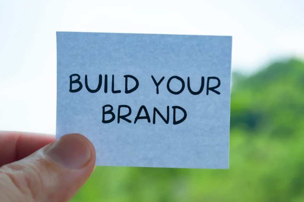 Eine Hand hält eine Karte mit dem Text „BUILD YOUR BRAND“ vor einem unscharfen grünen Hintergrund.