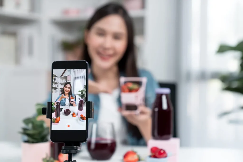 Eine Person filmt sich selbst, während sie einen Saftkarton hält. Die Kamera eines Smartphones ist auf sie gerichtet. Auf dem Tisch ist ein Aufbau mit Saftflaschen, einem Glas und Früchten zu sehen.
