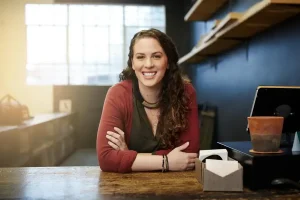 Eine lächelnde Frau mit verschränkten Armen sitzt an einem Schreibtisch in einer entspannten Büroumgebung.