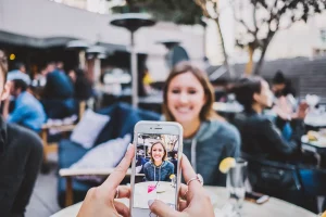 Person, die in einem Straßencafé ein Foto einer lächelnden Frau mit einem Smartphone macht.