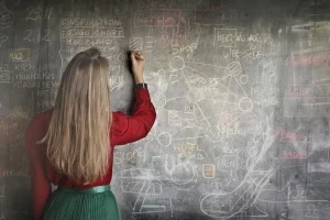 Eine Person mit langen Haaren schreibt auf eine Tafel, die mit verschiedenen Diagrammen und Notizen gefüllt ist. Sie steht mit dem Rücken zur Kamera und trägt ein rotes Hemd und einen grünen Rock.