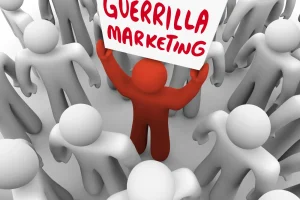 Eine rote Figur in einer Menschenmenge hält ein Schild mit der Aufschrift „Guerilla Marketing“ hoch. Die umliegenden Figuren sind grau.